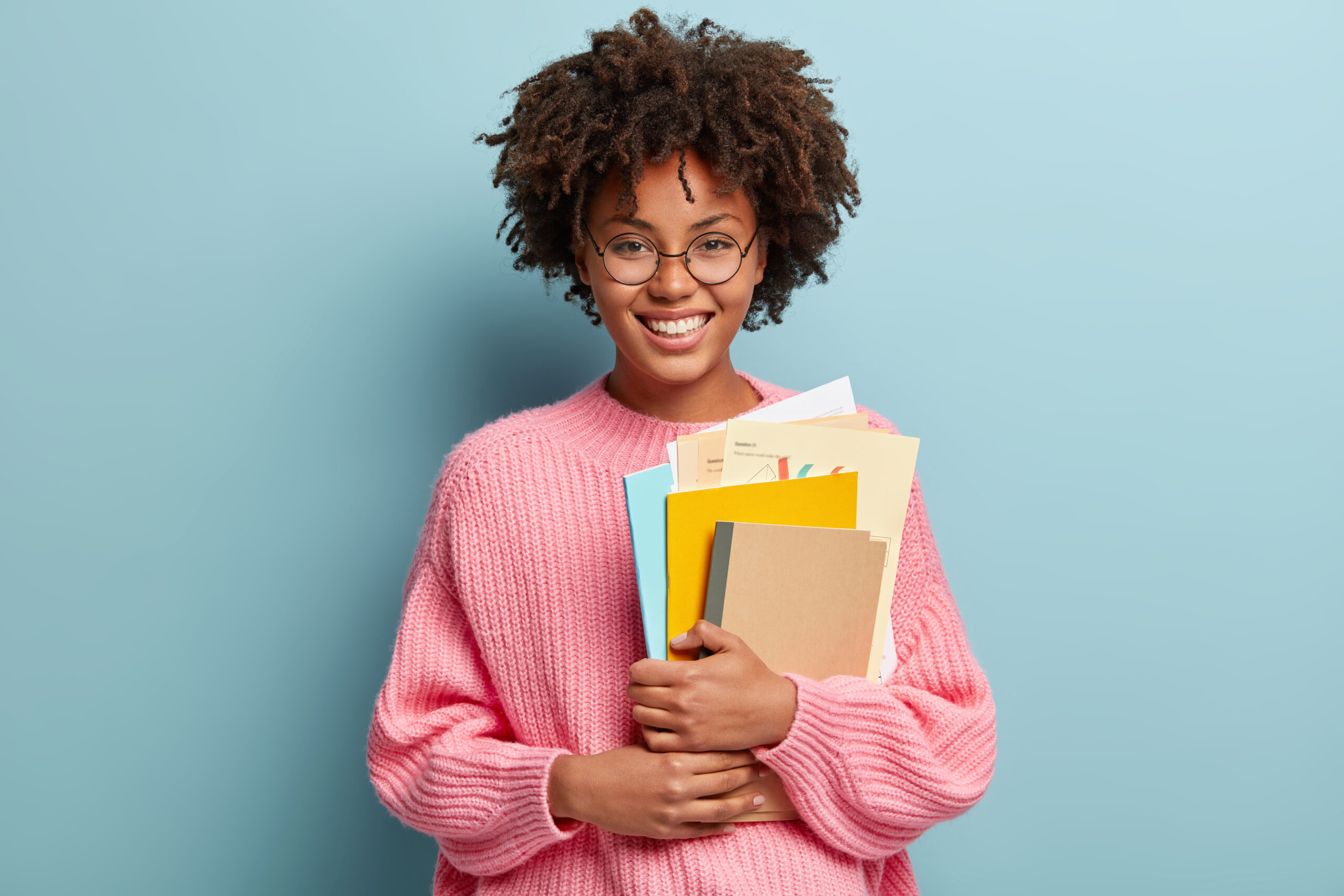 Education and ethnicity concept. Optimistic glad dark skinned young woman with pleased expression, holds textbooks nad papers, rejoices learning new information, isolated over blue background